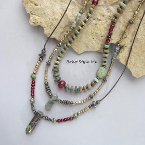 Puede incluir: Un collar de varias hebras con cuentas verdes y rojas, espaciadores met&aacute;licos y dos colgantes de piedra. El collar incluye elementos de cord&oacute;n marr&oacute;n y el texto Boho Style Me. El dise&ntilde;o incorpora varias formas y tama&ntilde;os de cuentas.