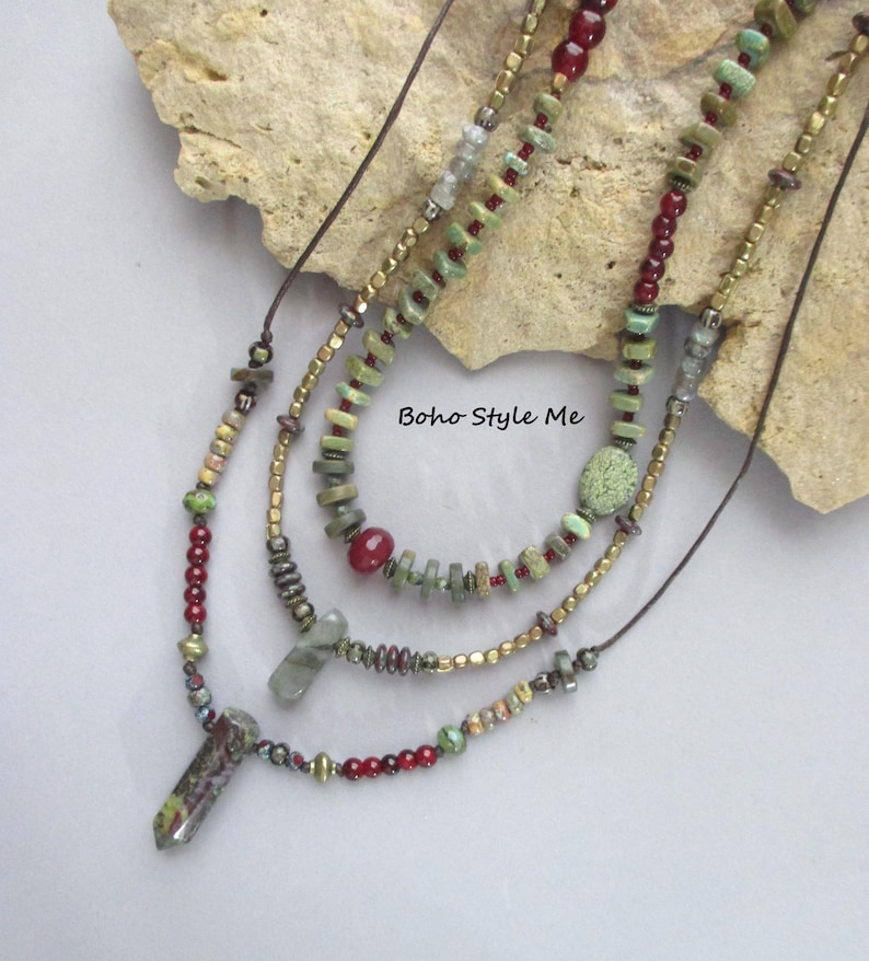 Puede incluir: Un collar de varias hebras con cuentas verdes y rojas, espaciadores met&aacute;licos y dos colgantes de piedra. El collar incluye elementos de cord&oacute;n marr&oacute;n y el texto Boho Style Me. El dise&ntilde;o incorpora varias formas y tama&ntilde;os de cuentas.