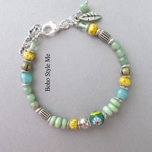 May include: A beaded bracelet featuring a mix of light green, turquoise, and yellow beads, interspersed with silver-toned spacers and a silver leaf charm. The bracelet includes a lobster claw clasp and the text Boho Style Me printed on the side.