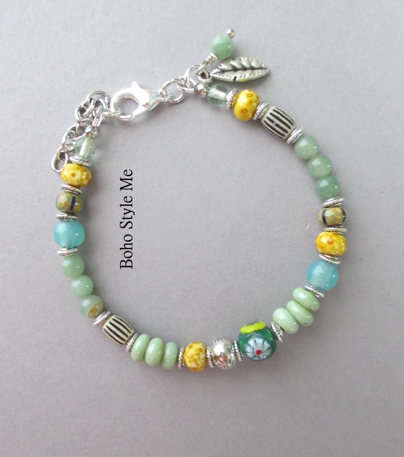 May include: A beaded bracelet featuring a mix of light green, turquoise, and yellow beads, interspersed with silver-toned spacers and a silver leaf charm. The bracelet includes a lobster claw clasp and the text Boho Style Me printed on the side.