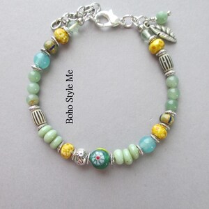 May include: A beaded bracelet featuring a mix of green, yellow, and blue beads, silver-toned spacers, and a central green bead with a red and white floral pattern. The bracelet includes a silver-toned lobster clasp and a small silver leaf charm.