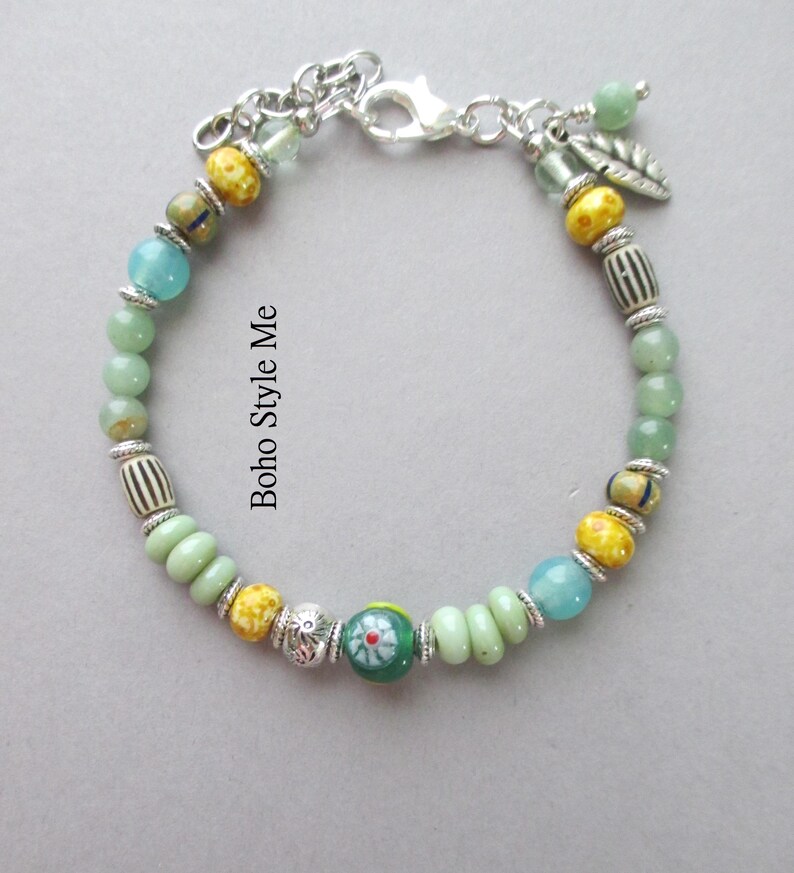 May include: A beaded bracelet featuring a mix of green, yellow, and blue beads, silver-toned spacers, and a central green bead with a red and white floral pattern. The bracelet includes a silver-toned lobster clasp and a small silver leaf charm.