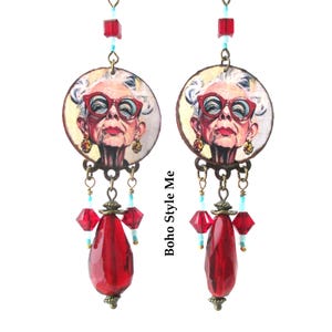May include: A pair of dangle earrings featuring circular pendants with an illustration of an older person wearing red glasses. The earrings include bronze-toned metal hardware, small light blue beads, red cube beads, and large faceted red teardrop beads.