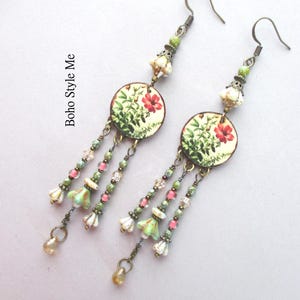 May include: A pair of dangle earrings featuring circular charms with botanical illustrations of red flowers and green foliage. The earrings include bronze-toned hooks, small green and pink beads, and bell-shaped floral accents. Text reads Boho Style Me.