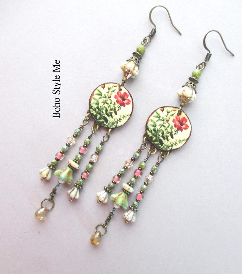 May include: A pair of dangle earrings featuring circular charms with botanical illustrations of red flowers and green foliage. The earrings include bronze-toned hooks, small green and pink beads, and bell-shaped floral accents. Text reads Boho Style Me.