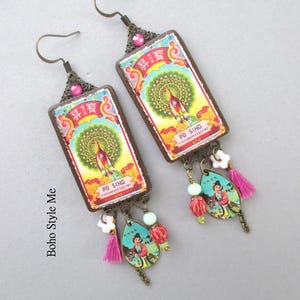 May include: A pair of rectangular drop earrings featuring a vintage-style label with a peacock illustration and the text PO SING. The earrings include bronze-tone metal accents, a pink crystal, a small white flower bead, a pink tassel, and a teardrop charm.