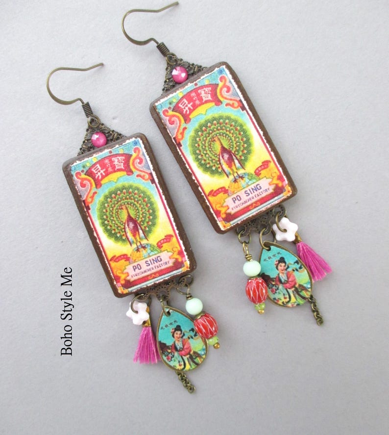 May include: A pair of rectangular drop earrings featuring a vintage-style label with a peacock illustration and the text PO SING. The earrings include bronze-tone metal accents, a pink crystal, a small white flower bead, a pink tassel, and a teardrop charm.