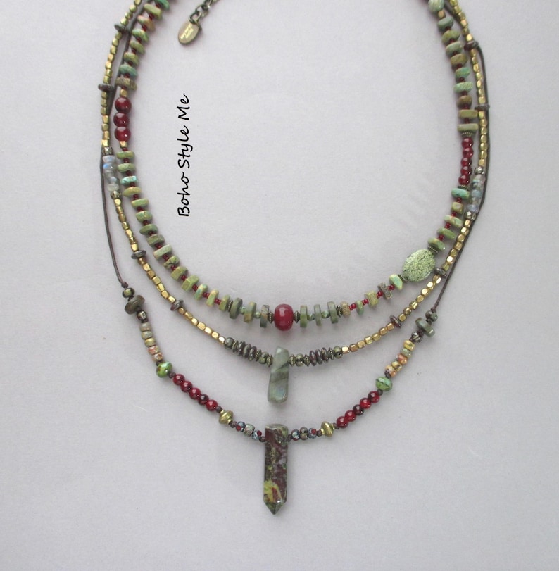 Puede incluir: Un collar de varias hebras con tres capas de cuentas en tonos verde, rojo y oro met&aacute;lico. El dise&ntilde;o incluye peque&ntilde;as cuentas en forma de disco, cuentas rojas redondas y dos colgantes de piedra. El texto Boho Style Me es visible.