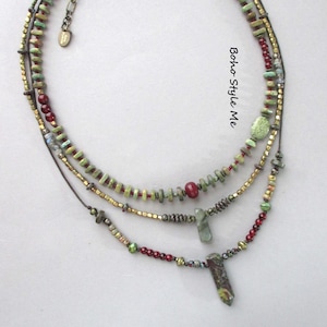 Puede incluir: Un collar de varias hileras con cuentas verdes, rojas y doradas met&aacute;licas, junto con dos colgantes de piedra. El collar incluye una peque&ntilde;a etiqueta met&aacute;lica con el texto Boho Style Me y presenta diversas formas y texturas de cuentas.
