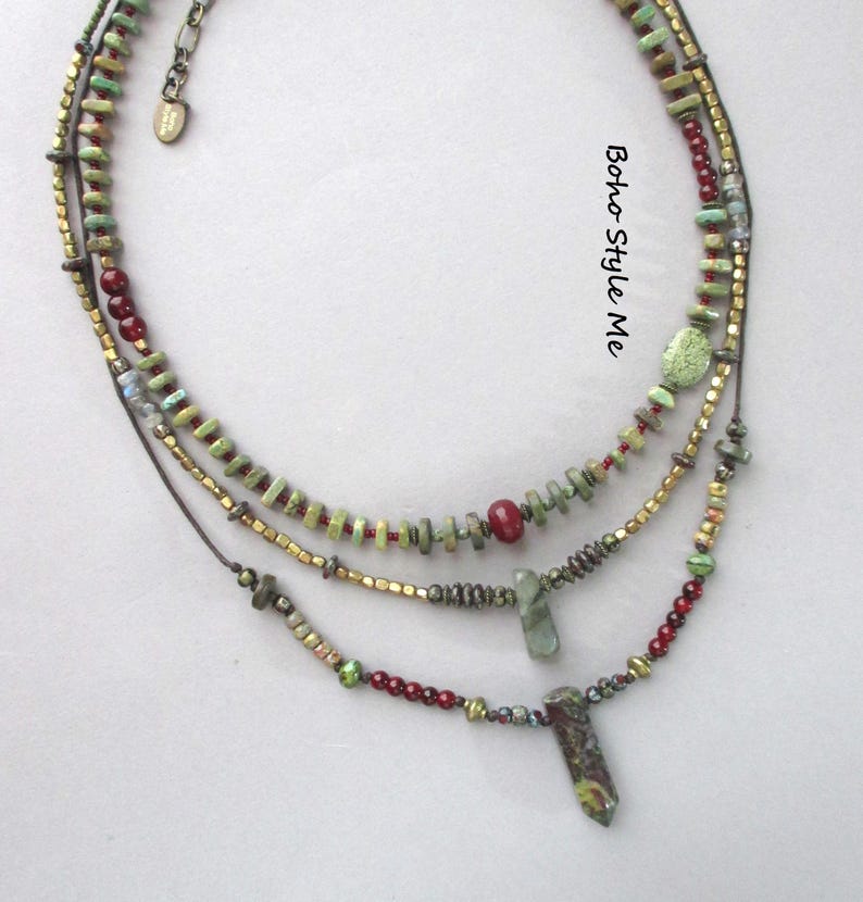 Puede incluir: Un collar de varias hileras con cuentas verdes, rojas y doradas met&aacute;licas, junto con dos colgantes de piedra. El collar incluye una peque&ntilde;a etiqueta met&aacute;lica con el texto Boho Style Me y presenta diversas formas y texturas de cuentas.