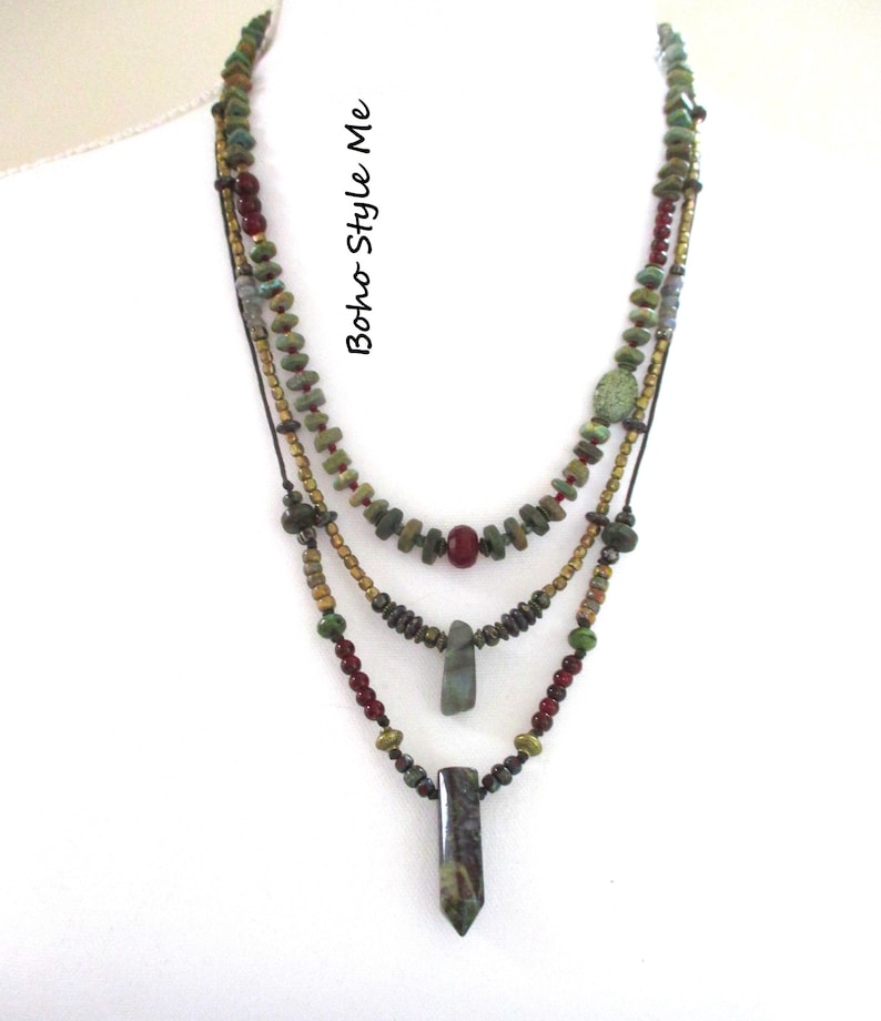 Puede incluir: Un collar de varias hebras con cuentas verdes, marrones y rojas y tres colgantes distintos. El texto "Boho Style Me" est&aacute; impreso verticalmente en el lateral. El dise&ntilde;o incorpora varias cuentas tipo piedra y acentos met&aacute;licos en capas.