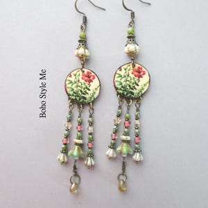 May include: A pair of dangle earrings featuring circular charms with a red floral print and the text Boho Style Me. The earrings include green, pink, and clear beads, along with small bell-shaped accents in white and green, suspended on metal hooks.