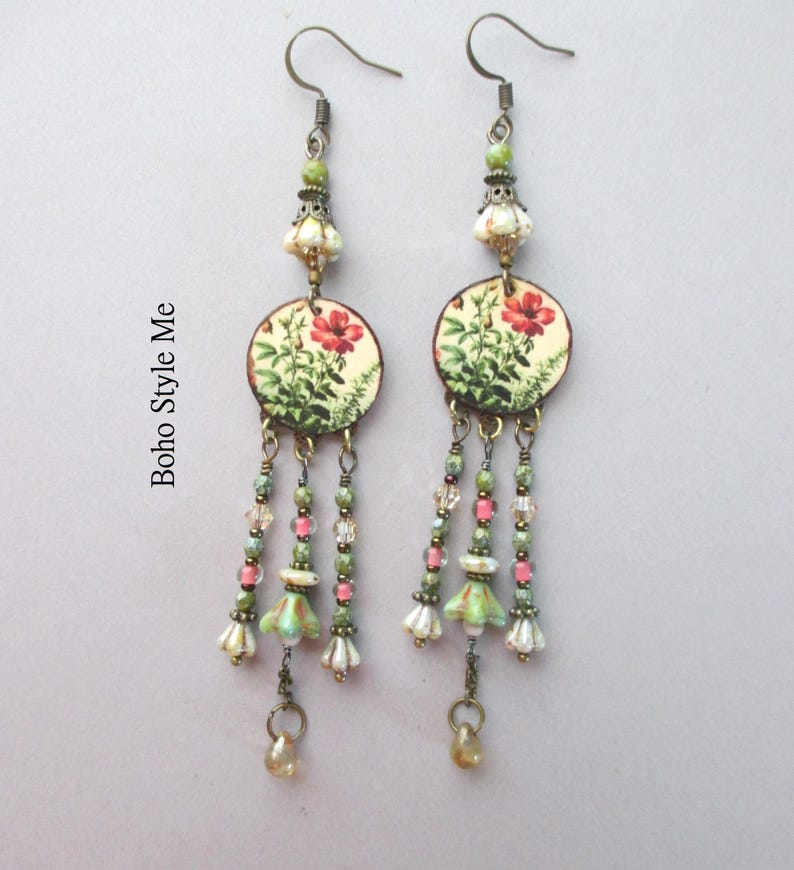 May include: A pair of dangle earrings featuring circular charms with a red floral print and the text Boho Style Me. The earrings include green, pink, and clear beads, along with small bell-shaped accents in white and green, suspended on metal hooks.