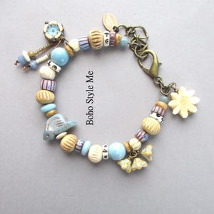 May include: A charm bracelet featuring a variety of beads in shades of light blue, cream, and tan, including a ceramic fish, a flower, and striped patterns. The bracelet includes a bronze-toned lobster clasp and the text Boho Style Me.