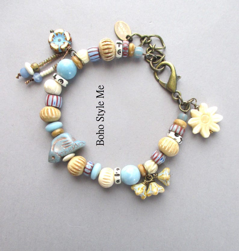 May include: A charm bracelet featuring a variety of beads in shades of light blue, cream, and tan, including a ceramic fish, a flower, and striped patterns. The bracelet includes a bronze-toned lobster clasp and the text Boho Style Me.