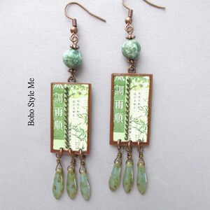 Puede incluir: Un par de pendientes colgantes con ganchos de tono cobre, cuentas redondas de color verde moteado y colgantes rectangulares. Los colgantes muestran papel verde con ilustraciones de bamb&uacute;, texto japon&eacute;s y una l&iacute;nea vertical de cuentas. Tres peque&ntilde;as cuentas verdes en forma de l&aacute;grima cuelgan de la base de cada marco rectangular.