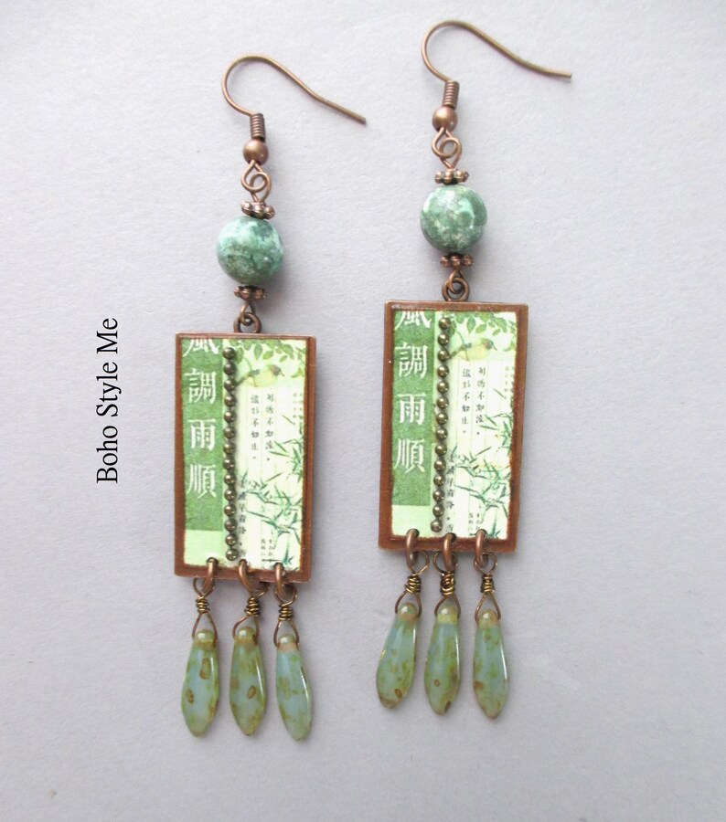 Puede incluir: Un par de pendientes colgantes con ganchos de tono cobre, cuentas redondas de color verde moteado y colgantes rectangulares. Los colgantes muestran papel verde con ilustraciones de bamb&uacute;, texto japon&eacute;s y una l&iacute;nea vertical de cuentas. Tres peque&ntilde;as cuentas verdes en forma de l&aacute;grima cuelgan de la base de cada marco rectangular.