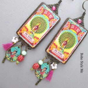 May include: A pair of rectangular drop earrings featuring a vintage-style label with a peacock illustration and the text PO SING FIRECRACKER FACTORY. Each earring includes a teardrop charm with a figure, a pink tassel, and colourful beads.