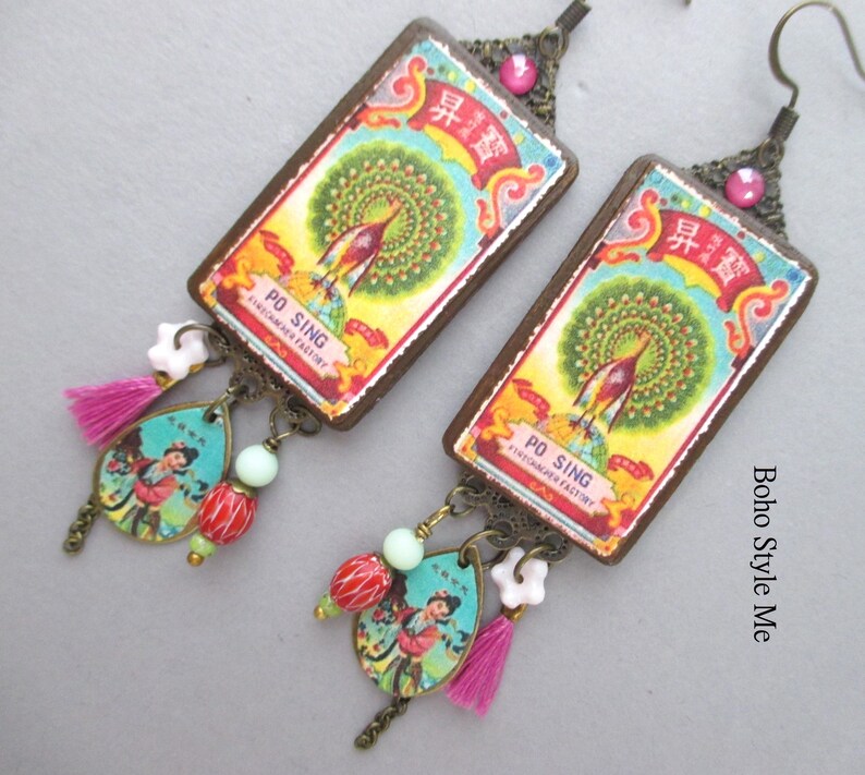 May include: A pair of rectangular drop earrings featuring a vintage-style label with a peacock illustration and the text PO SING FIRECRACKER FACTORY. Each earring includes a teardrop charm with a figure, a pink tassel, and colourful beads.