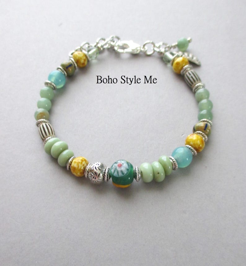 May include: A beaded bracelet featuring a mix of light green, yellow, and blue beads interspersed with silver-toned metal spacers and a central green bead with a floral pattern. The text Boho Style Me is visible. It includes a silver-toned clasp.