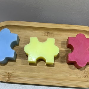 Puede incluir: Tres piezas de jabón con forma de rompecabezas en una bandeja de madera. El jabón es azul, amarillo y rosa.