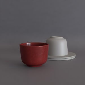 Può includere: Una tazza in ceramica rosso scuro con una consistenza maculata posizionata davanti a una tazza grigio chiaro opaca e liscia capovolta sul suo piattino abbinato. La tazza rossa ha una base arrotondata e un'apertura ampia, mentre quella grigia è semplice.