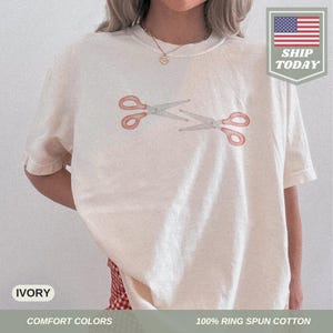 May include: Cream-coloured t-shirt featuring a watercolour design of two scissors. The scissors have red handles and grey blades. The shirt is made of 100% ring-spun cotton and is part of the comfort colours collection.