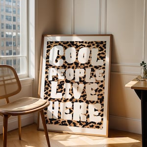 Peut inclure: Une affiche encadrée avec le texte "COOL PEOPLE LIVE HERE" en lettres blanches sur un fond imprimé léopard. L'œuvre est exposée dans une pièce avec une chaise en bois et une petite table.