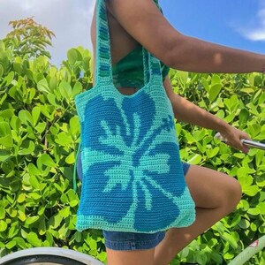 May include: A crocheted tote bag with a blue and green hibiscus flower design. The bag has a long strap and is worn over the shoulder.