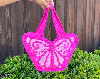 May include: A pink crocheted butterfly-shaped handbag with white accents. The bag has two straps and is held by a hand.