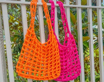 May include: Two crocheted mesh bags hanging on a white fence. One bag is orange and the other is pink.
