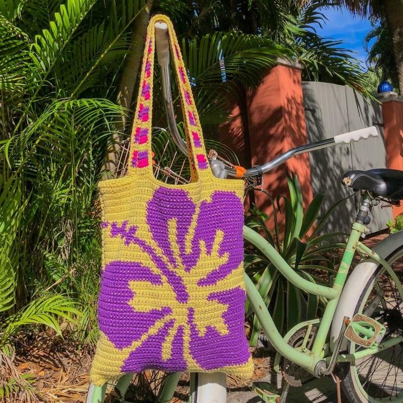 May include: A yellow crocheted tote bag with a purple hibiscus flower design. The bag is hanging on a bicycle.