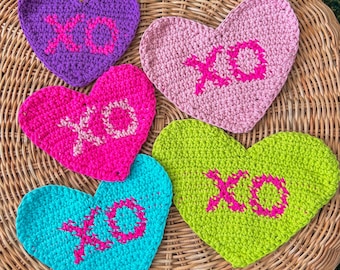 May include: Five crocheted heart-shaped coasters with the letters XO in pink on a light brown wicker surface.