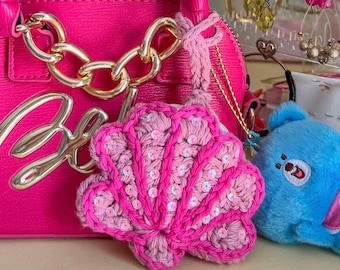 May include: A bright pink handbag with gold chain detailing and a crocheted seashell charm. The charm is pink with sequins. A small blue plush toy is visible in the background.