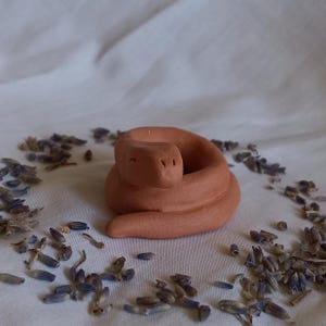 May include: A coiled terracotta snake figurine with a smiling face. The handmade ceramic sculpture is a warm, earthy tone. Dried lavender buds are scattered around the snake on a white cloth, creating a natural, rustic aesthetic.