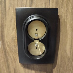 May include: A black electrical outlet cover with two round openings. The openings are made of a light brown material.