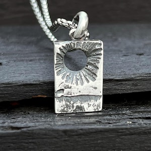 May include: A silver pendant necklace. The rectangular pendant has a textured surface and a circular opening. The pendant hangs from a delicate silver chain. The necklace is shown on a dark, textured surface.
