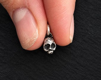 May include: A small, three-dimensional silver-toned skull charm featuring a weathered, textured surface with deep-set eye sockets and a nasal cavity. The charm includes a small circular jump ring at the top for attachment to a chain or bracelet.