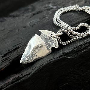 Könnte beinhalten: Halskette mit einem silbernen Pfeilspitzen-Anhänger. Der Anhänger hat ein strukturiertes, verwittertes Aussehen und hängt an einer filigranen Kette. Die Halskette wird vor einem dunklen, strukturierten Hintergrund präsentiert.