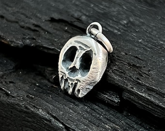May include: A small, silver-colored skull pendant with large eye sockets and a detailed jawline. The pendant has a loop at the top for a chain. The skull is set against a dark, textured background.