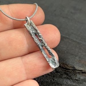 Könnte beinhalten: Halskette mit silbernem Anhänger. Der rechteckige Anhänger hat eine strukturierte Oberfläche und ein ausgeschnittenes Design. Er hängt an einer feinen Silberkette. Die Halskette wird vor dunklem Hintergrund gezeigt.