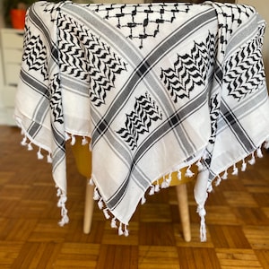 Könnte beinhalten: Ein weiß-schwarzer Keffiyeh-Schal mit geometrischem Muster und Fransenbesatz. Der Schal ist über einen Stuhl drapiert und zeigt sein Design. Der Schal hat ein traditionelles Design mit schwarz-weißen Mustern.