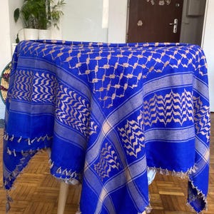 Könnte beinhalten: Ein leuchtend blauer Schal mit einem beigefarbenen geometrischen Muster. Der Schal ist über einem Tisch drapiert und zeigt sein aufwendiges Design und die gefransten Kanten. Der Stoff wirkt weich und leicht, geeignet für verschiedene Zwecke.