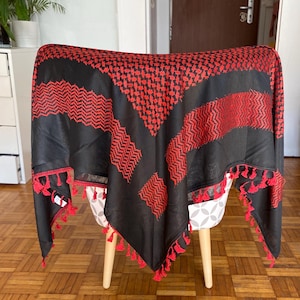 Könnte beinhalten: Schwarzer und roter Keffiyeh-Schal mit geometrischen Mustern und roter Quastenborte. Der Schal ist über einem Stuhl drapiert. Der Keffiyeh ist eine traditionelle Kopfbedeckung aus dem Nahen Osten, oft als Schal getragen.