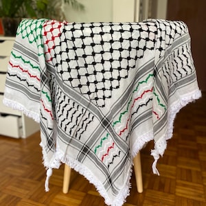 Könnte beinhalten: Ein weißes Keffiyeh-Tuch mit einem geometrischen Muster in Schwarz, Rot und Grün. Das Tuch ist über einen Holzstuhl drapiert und hat gefranste Kanten. Das Design umfasst ein Gittermuster und Zickzacklinien.