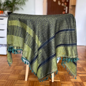 Könnte beinhalten: Ein grün-schwarz gemusterter Schal mit Zickzack-Design. Der Schal hat einen Fransenbesatz aus türkisfarbenen Quasten an den Kanten. Der Schal ist über einen Holzstuhl drapiert.