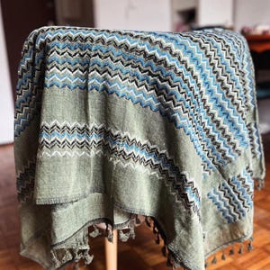 Könnte beinhalten: Eine gemusterte Decke in Grün-, Blau- und Grautönen, die über einem kleinen Tisch drapiert ist. Die Decke hat ein Zickzack-Muster und Fransen an den Kanten. Der Tisch hat eine helle Holzoberfläche.