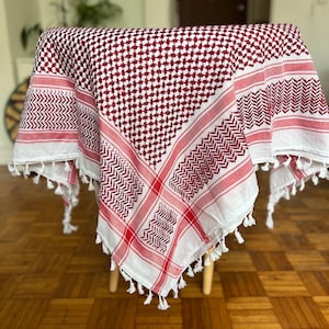 Könnte beinhalten: Ein rot-weiß kariertes Keffiyeh-Tuch, das über einem kleinen Tisch drapiert ist. Der Schal hat ein sich wiederholendes Muster aus kleinen Quadraten und dekorativen Bordüren mit Quasten an den Kanten. Der Schal ist aus weichem Stoff.