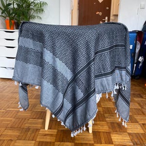 Könnte beinhalten: Ein grau-schwarzer Schal mit Muster und weißen Quasten. Der Schal ist über einen kleinen Holzstuhl drapiert. Das Muster umfasst ein Zickzack-Design und Streifen. Der Schal scheint aus einem weichen Stoff zu sein.