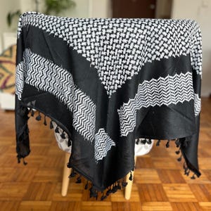 Könnte beinhalten: Schwarz-weißer Keffiyeh-Schal mit geometrischem Muster und Quastenbesatz. Der Schal ist über einem Stuhl drapiert, um sein Design zu zeigen. Das Muster umfasst sich wiederholende Quadrate und diagonale Linien.
